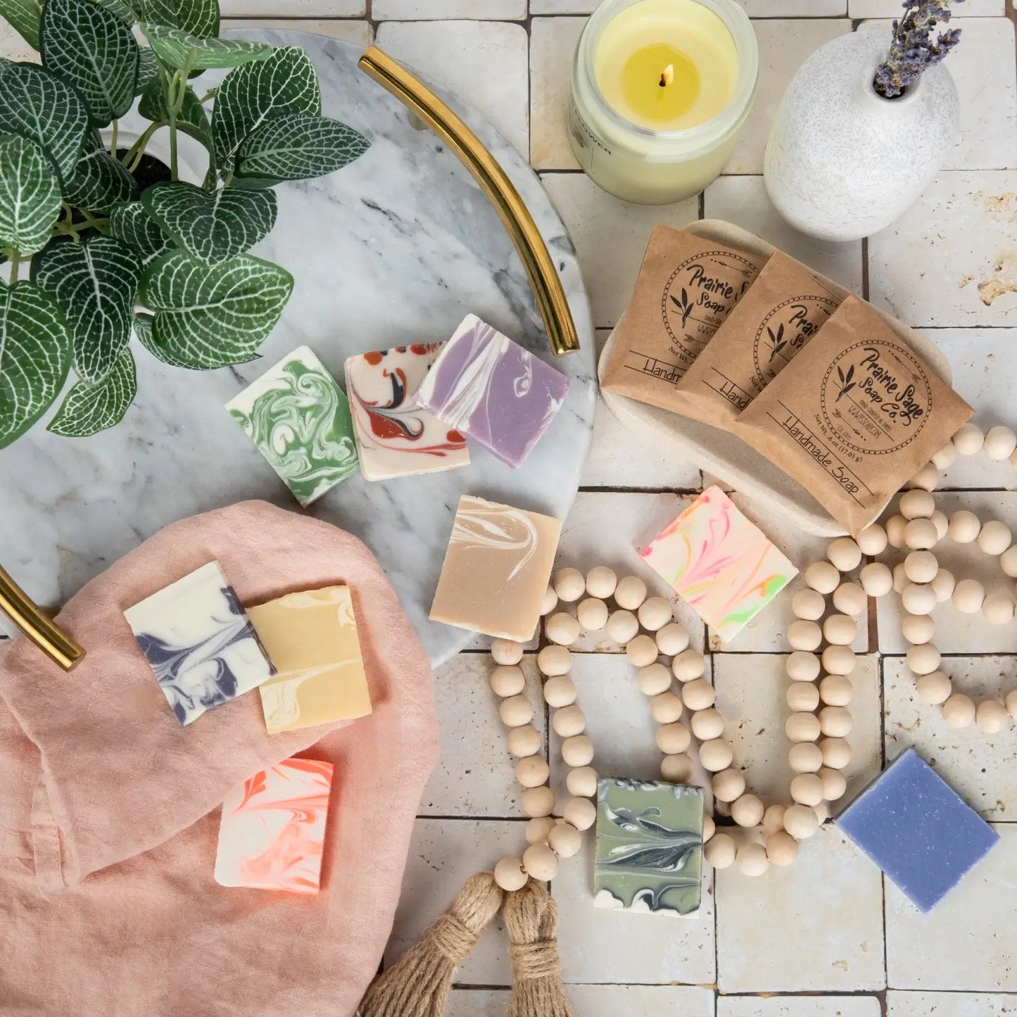 Handmade sample soap bars with natural packaging on a wooden surface with a candle and plant.