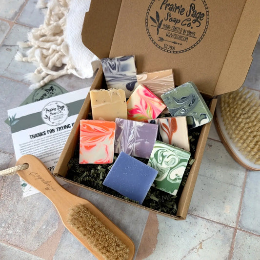 Box of assorted soaps with a brush and leafy branch on a tiled surface, featuring Prairie Sage Soap Co branding.