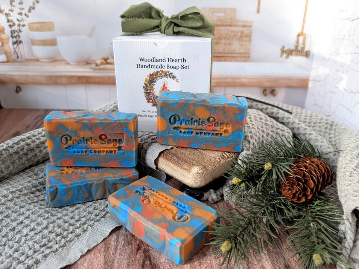 Our Indian Summer scented handmade soap displayed on a rustic wooden surface with a knitted blanket, pine branches, a pinecone, and a branded white box behind the bars. The soap bars are colored blue and orange with the brand name Prairie Sage Soap Company visible.