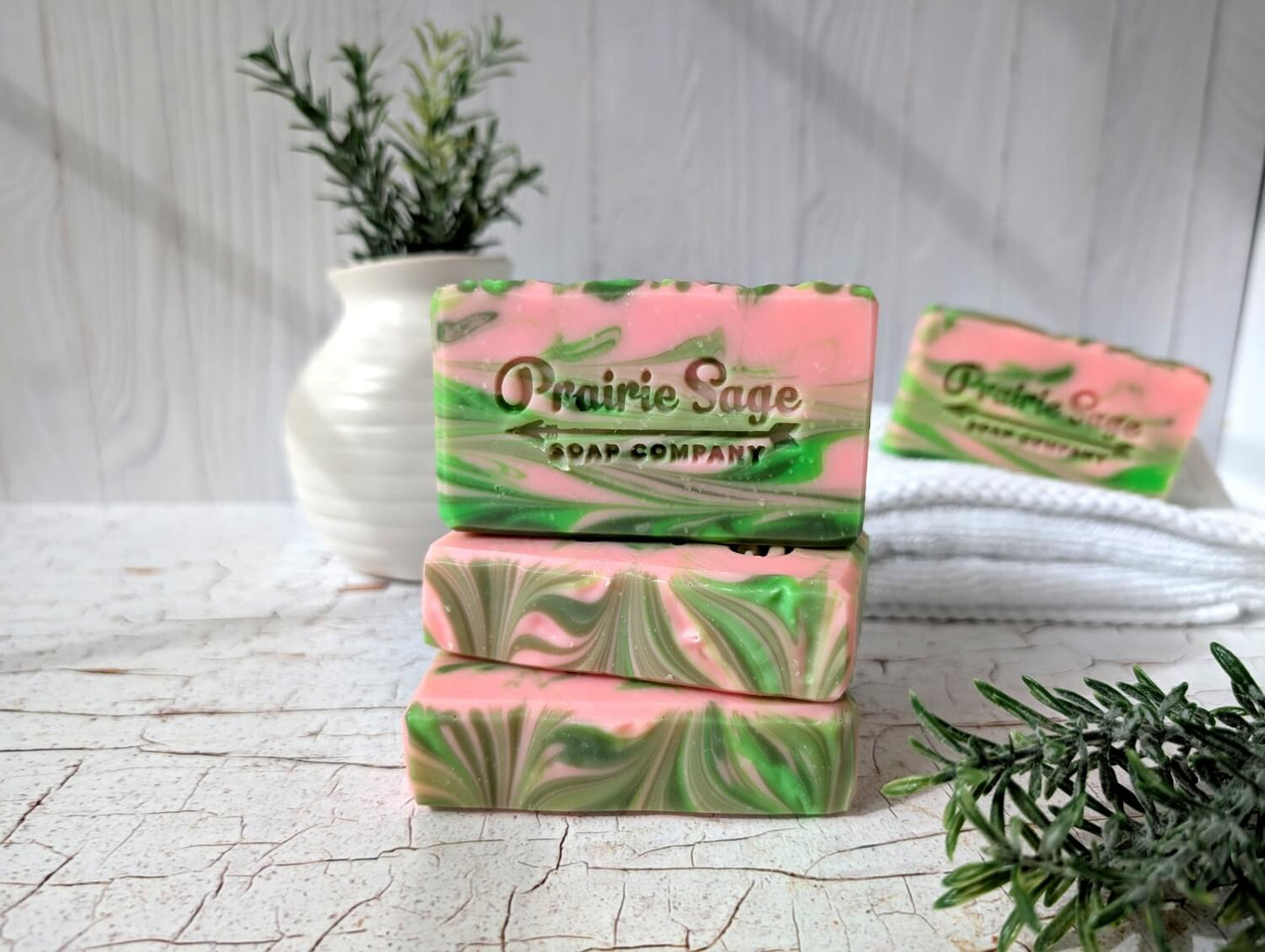 Stack of pink and green marbled soap bars with 'Prairie Sage Soap Company' branding on a light wooden surface.