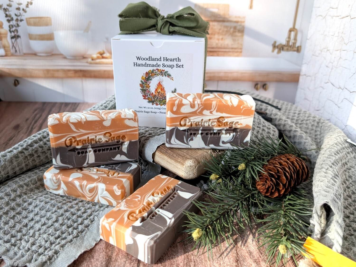 A set of Toasted Chestnut and Spice scented handmade soap bars from Prairie Sage Soap Company arranged on a textured gray blanket with pine branches, a pinecone, and a white gift box in the background.