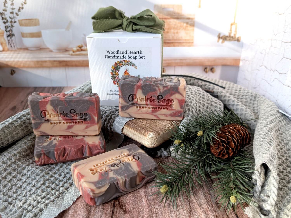 A set of Twigs and Berries scented handmade soap bars arranged with a white gift box tied with an olive green ribbon, pine branches, a pinecone, and a textured blanket on a wooden surface.