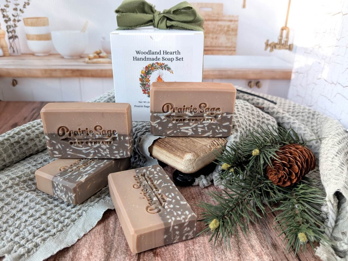 A set of White Pumpkin and Amber scented handmade soap bars from Prairie Sage Handmade Soap Co. arranged on a rustic wooden surface with a knitted blanket, pine branches, a pinecone, and the branded gift box in the background.