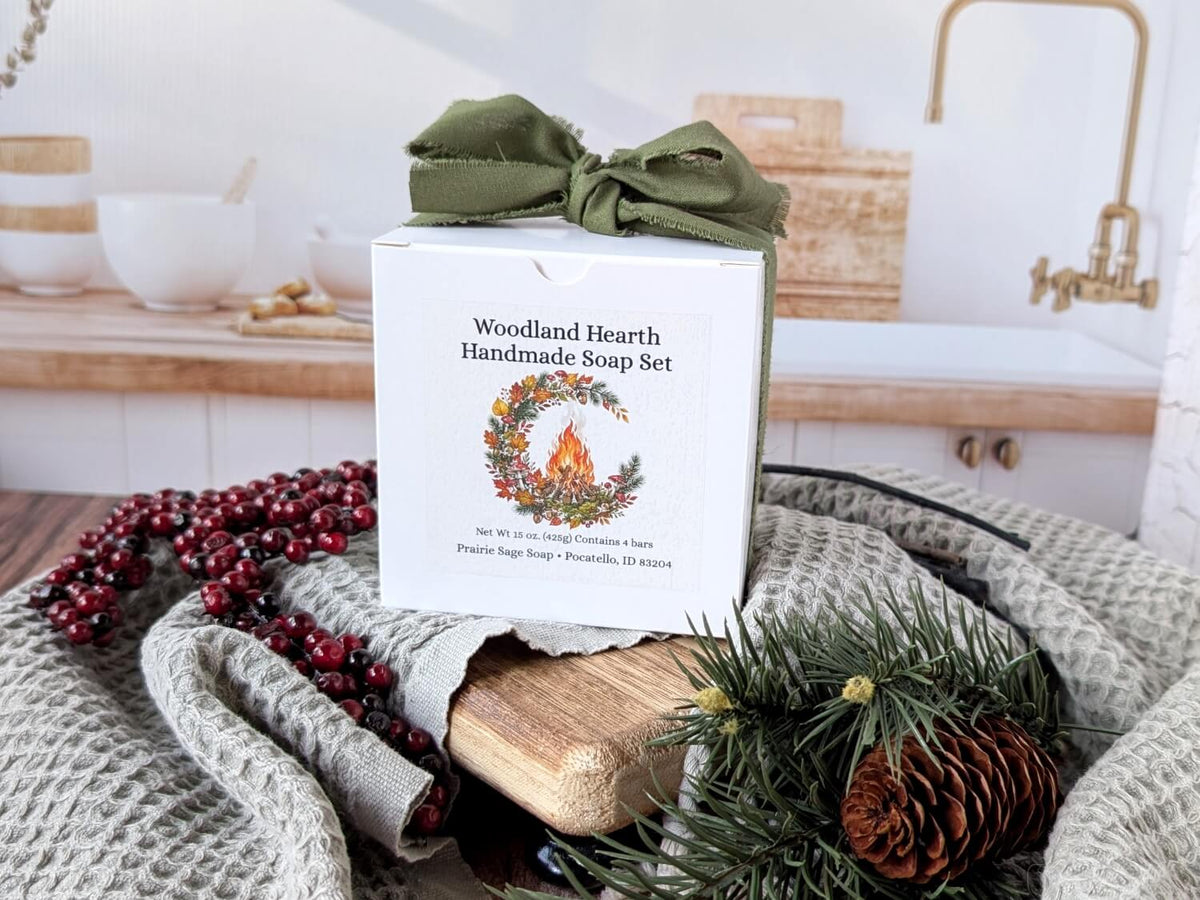 Woodland Hearth Handmade Soup Set in a white gift box with an olive green ribbon, styled on a gray knit blanket with pine branches, a pinecone, red berries, and a wooden block in a warm kitchen setting.