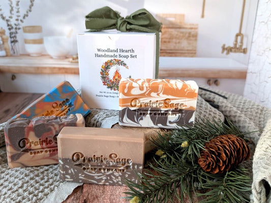 A gift set of Prairie Sage Handmade Soap bars and a branded box arranged on a knitted blanket with pine branches and a pinecone. The set includes a white box labeled 'Woodland Hearth Handmade Soap Set' and four soap bars in earthy tones: orange and cream, brown and cream, and multicolor with blue, orange, and brown.